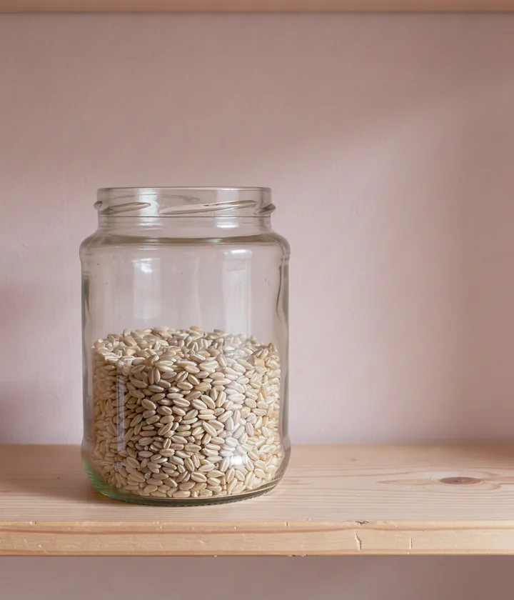 A plain glass jar of barley on a pale wooden pantry shelf